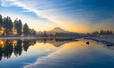 Sunrise over a calm lake with a snow-capped mountain and pine forest reflected in the water.