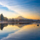Sunrise over a calm lake with a snow-capped mountain and pine forest reflected in the water.