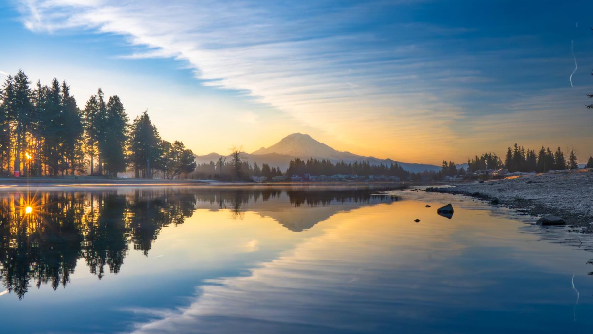 Sunrise over a calm lake with a snow-capped mountain and pine forest reflected in the water.
