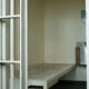 Prison cell interior with white metal bars and a perforated metal bed frame inside a small cell