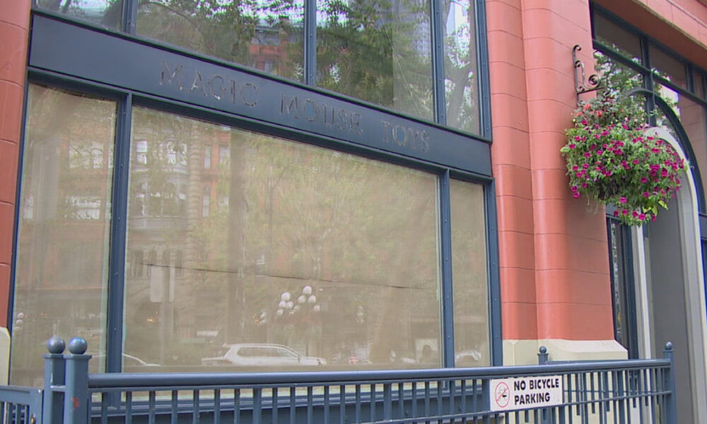 Front of a storefront with large display windows, pink-orange brick façade, and a hanging flower basket; a blue railing runs along the sidewalk with a 'No Bicycle Parking' sign visible.