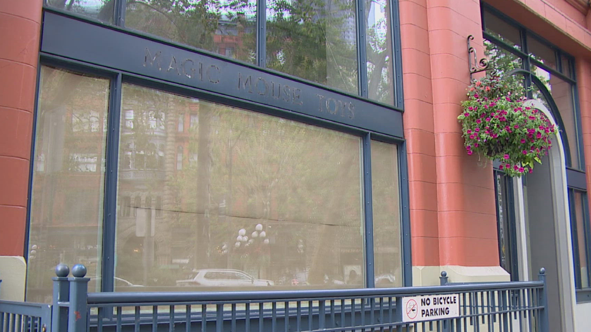 Front of a storefront with large display windows, pink-orange brick façade, and a hanging flower basket; a blue railing runs along the sidewalk with a 'No Bicycle Parking' sign visible.