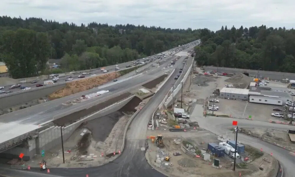 Aerial view of a highway under construction with an elevated roadway, heavy equipment, and orange barriers, surrounded by trees and construction trailers.
