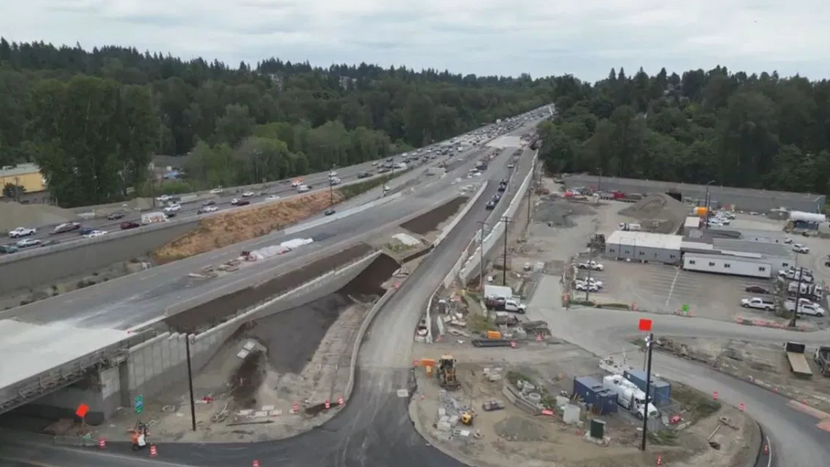 Aerial view of a highway under construction with an elevated roadway, heavy equipment, and orange barriers, surrounded by trees and construction trailers.