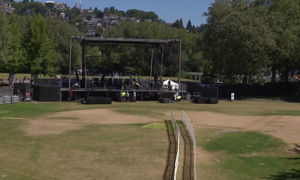 Outdoor concert stage set up in a park with rigging, speakers, and crew preparing on and around the stage; a grassy field in the foreground and trees in the background.