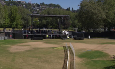 Outdoor concert stage set up in a park with rigging, speakers, and crew preparing on and around the stage; a grassy field in the foreground and trees in the background.