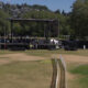 Outdoor concert stage set up in a park with rigging, speakers, and crew preparing on and around the stage; a grassy field in the foreground and trees in the background.