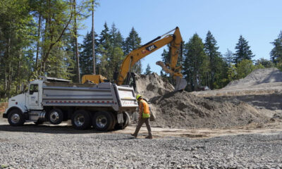 Excavator loading dirt into a white dump truck at a forested construction site; worker in an orange safety vest nearby.