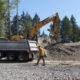Excavator loading dirt into a white dump truck at a forested construction site; worker in an orange safety vest nearby.