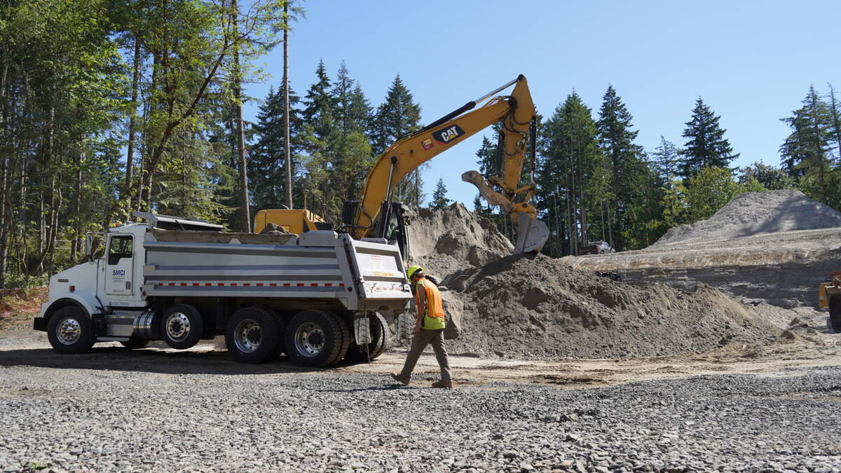Excavator loading dirt into a white dump truck at a forested construction site; worker in an orange safety vest nearby.