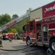 Firefighters battle a blaze near a red storefront; a tall ladder extends from a fire truck toward the building.