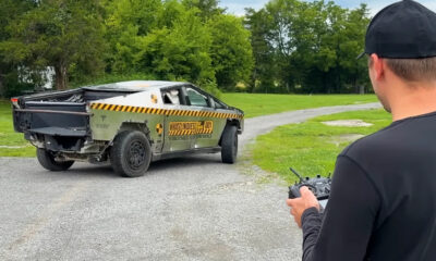 Man in a black cap drives an olive-drab armored pickup with yellow caution stripes and 'WhistlinDiesel' branding on a gravel road.