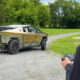 Man in a black cap drives an olive-drab armored pickup with yellow caution stripes and 'WhistlinDiesel' branding on a gravel road.