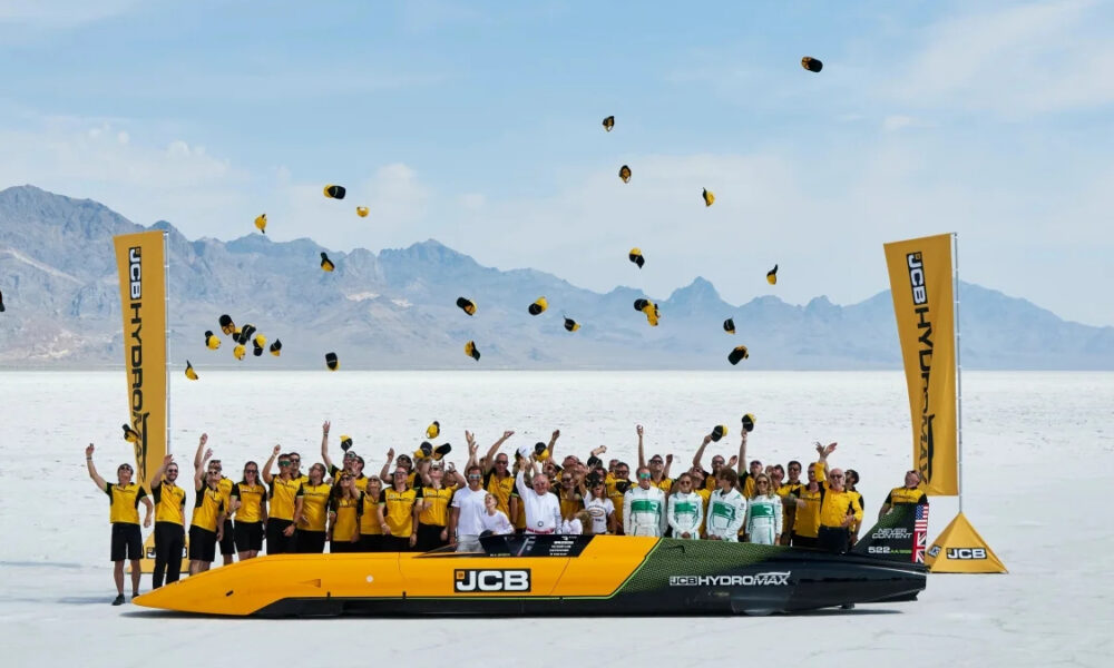 Team in yellow shirts on a bright yellow JCB hydro race boat on a salt flat, tossing hats into the air with mountains in the background.