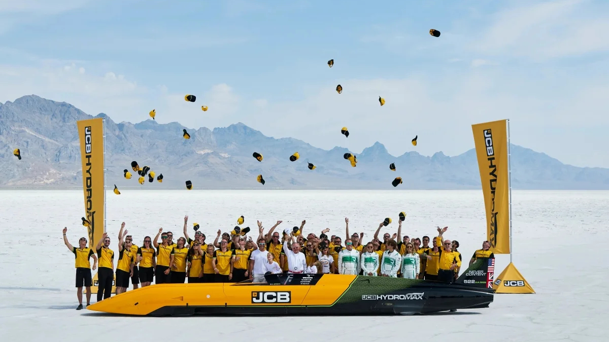 Team in yellow shirts on a bright yellow JCB hydro race boat on a salt flat, tossing hats into the air with mountains in the background.