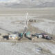 Oil drilling rig setup in a snowy desert with support trailers and multiple pickup trucks nearby. A remote field site under mountains in winter.