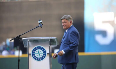 Man in a blue suit standing at a podium with a Seattle Mariners logo, speaking to a crowd at a stadium.