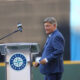 Man in a blue suit standing at a podium with a Seattle Mariners logo, speaking to a crowd at a stadium.