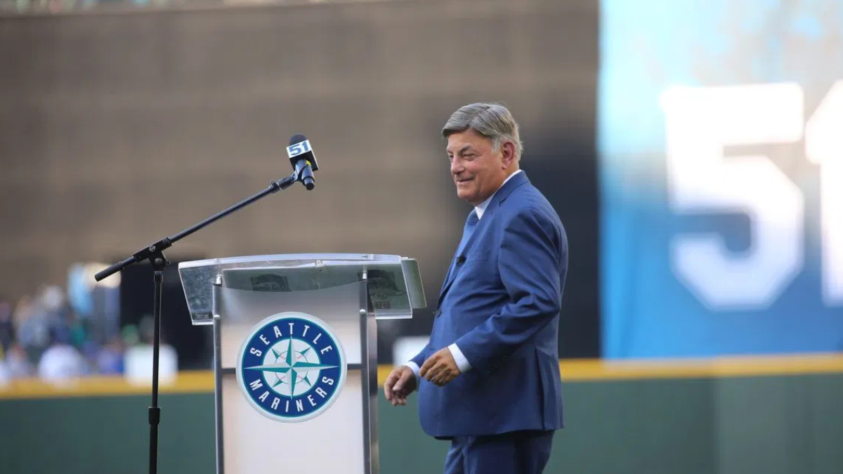 Man in a blue suit standing at a podium with a Seattle Mariners logo, speaking to a crowd at a stadium.