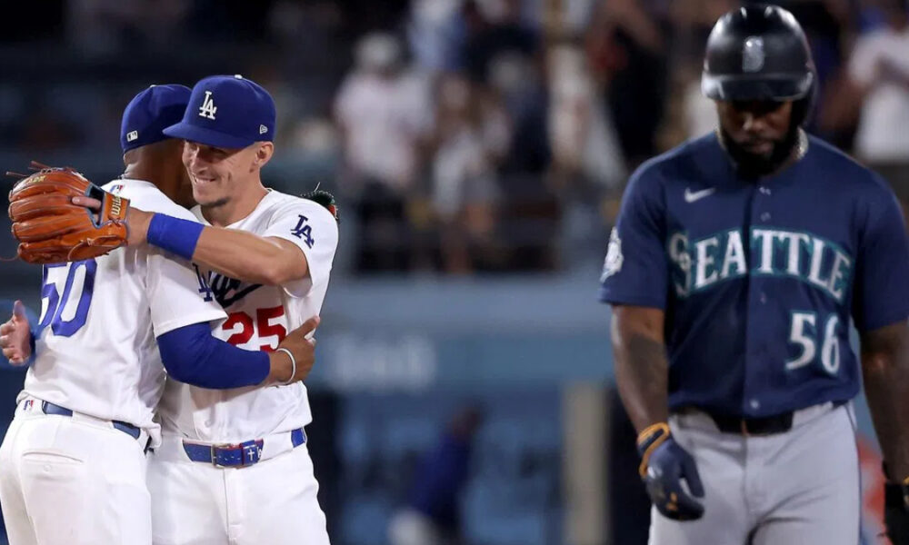 Two Los Angeles Dodgers players hug in celebration on the field as a Seattle Mariners player walks away in the foreground.