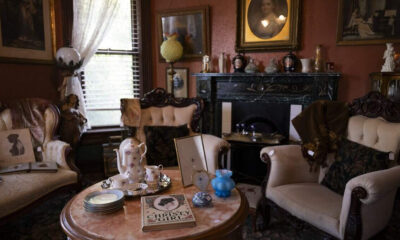 Vintage Victorian living room with tufted chairs, marble fireplace, and a tea set on a round table; framed portraits and decorative items fill the shelves.