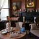 Vintage Victorian living room with tufted chairs, marble fireplace, and a tea set on a round table; framed portraits and decorative items fill the shelves.