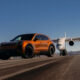 Orange SUV on an airport runway with a large white jet in the background under a clear blue sky.