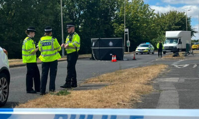 Police officers in high-visibility jackets stand on a cordoned road near a dark tent and a white van at an incident scene.