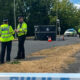 Police officers in high-visibility jackets stand on a cordoned road near a dark tent and a white van at an incident scene.