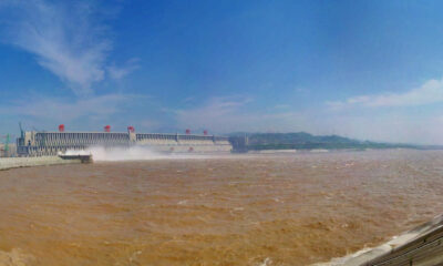 Hydroelectric dam along a muddy brown river, water spraying from spillways under a blue sky.