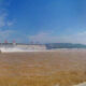 Hydroelectric dam along a muddy brown river, water spraying from spillways under a blue sky.