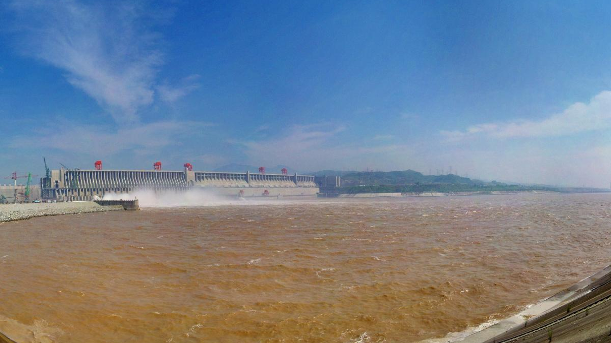 Hydroelectric dam along a muddy brown river, water spraying from spillways under a blue sky.
