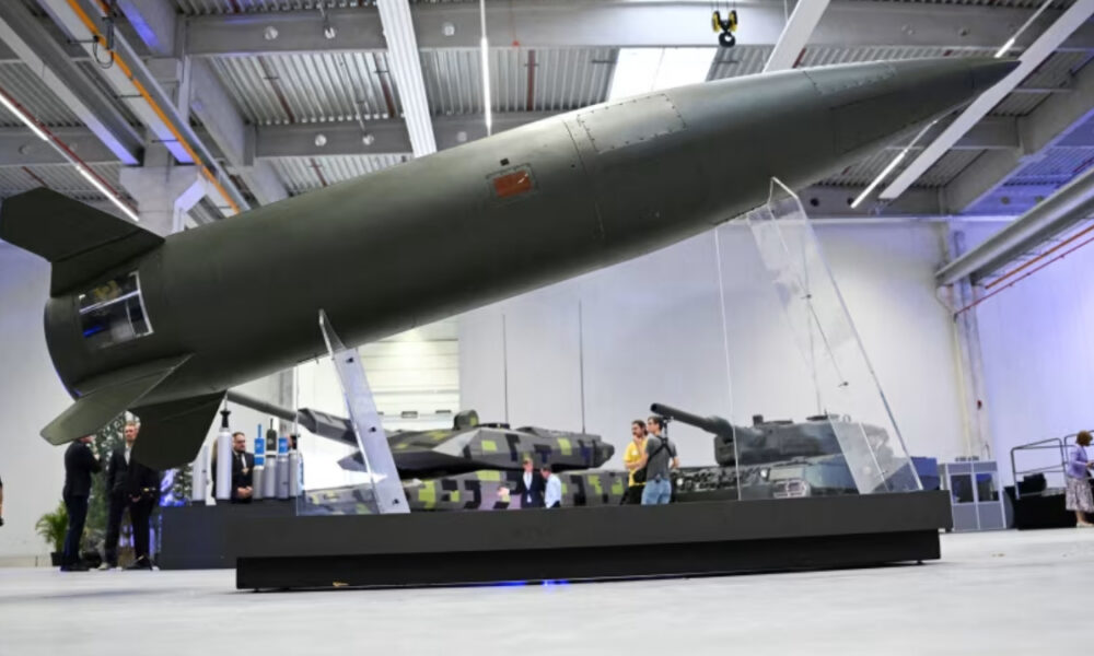 Large military missile displayed indoors on a raised platform, with armored vehicles visible in the background Exhibition hall setting with visitors observing the exhibit
