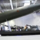 Large military missile displayed indoors on a raised platform, with armored vehicles visible in the background Exhibition hall setting with visitors observing the exhibit