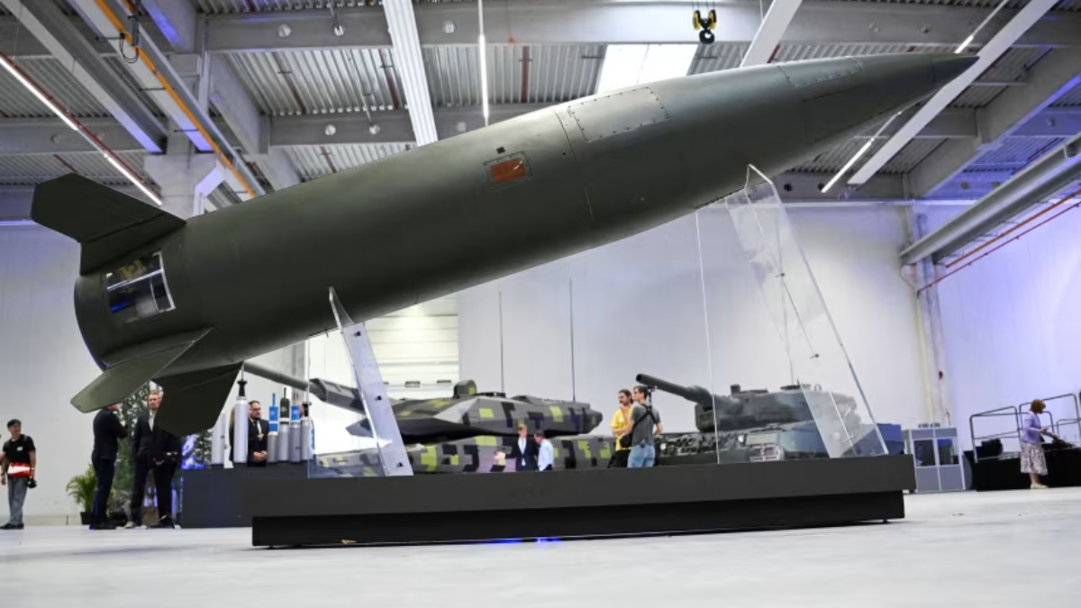 Large military missile displayed indoors on a raised platform, with armored vehicles visible in the background Exhibition hall setting with visitors observing the exhibit