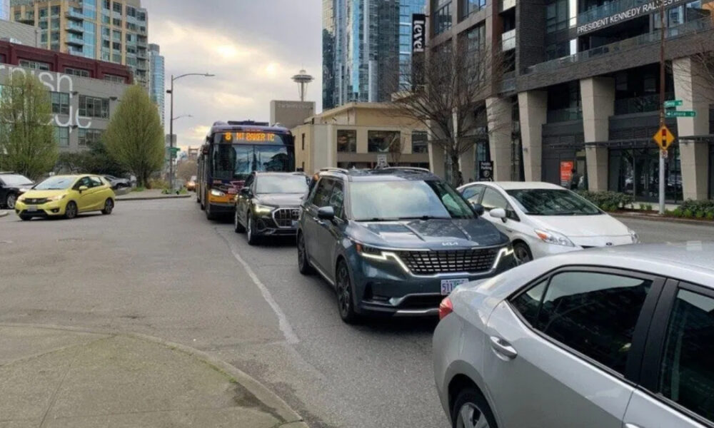 Bus and several cars stuck in traffic on a city street with tall modern buildings in the background and a yellow car on the left edge.