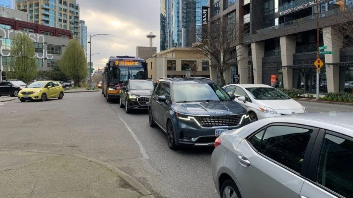 Bus and several cars stuck in traffic on a city street with tall modern buildings in the background and a yellow car on the left edge.