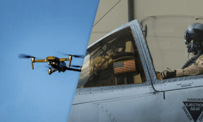 A small drone flies beside a gray military fighter jet cockpit with a pilot inside wearing a helmet and oxygen mask, near an American flag window.