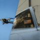 A small drone flies beside a gray military fighter jet cockpit with a pilot inside wearing a helmet and oxygen mask, near an American flag window.