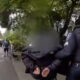 Police officer detains a person with hands behind their back during a street festival scene, bystanders and tents in the background.