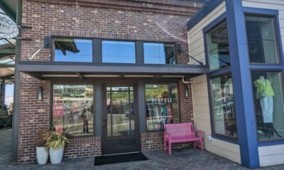 Brick storefront with glass doors, large windows, pink bench outside, and potted plants; clothing displays visible inside.