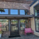 Brick storefront with glass doors, large windows, pink bench outside, and potted plants; clothing displays visible inside.