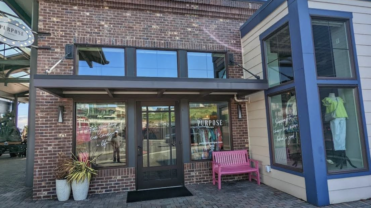 Brick storefront with glass doors, large windows, pink bench outside, and potted plants; clothing displays visible inside.