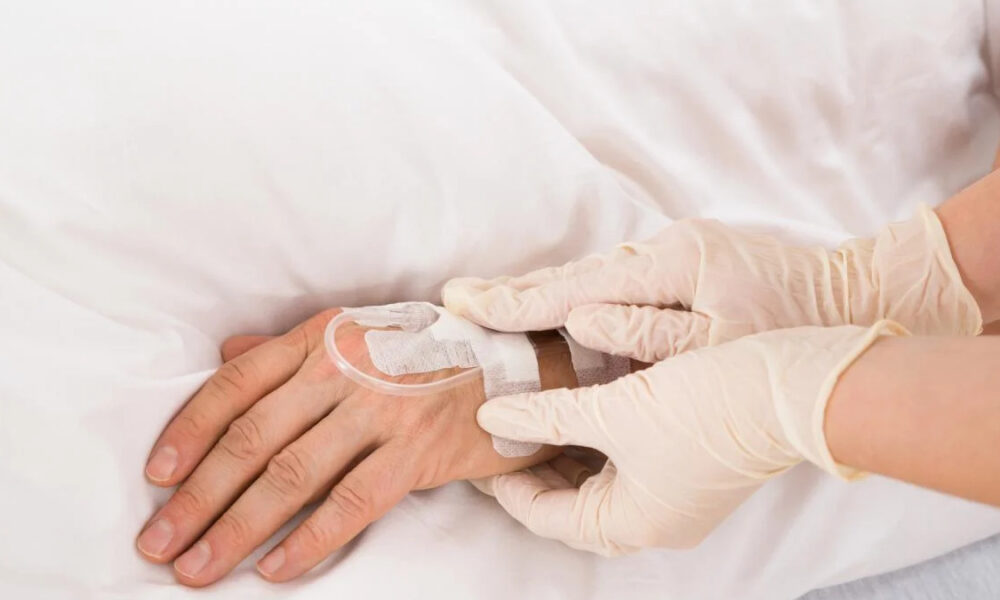 Healthcare worker in gloves securing an IV line on a patient’s hand with tape and tubing visible