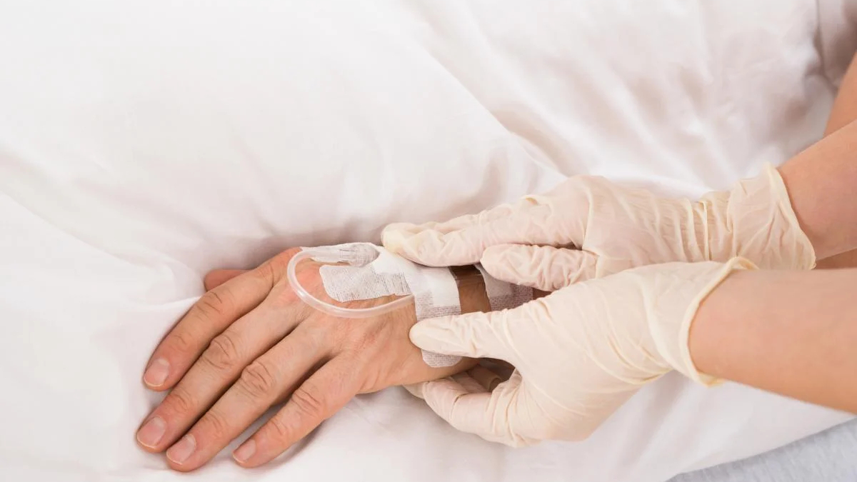 Healthcare worker in gloves securing an IV line on a patient’s hand with tape and tubing visible