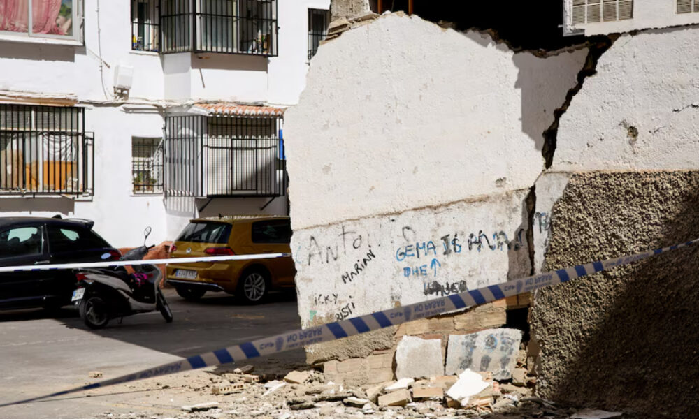 Crumbled concrete wall with rubble on a street, police tape across, and parked cars and a scooter in front of white apartment buildings.