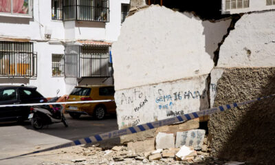 Crumbled concrete wall with rubble on a street, police tape across, and parked cars and a scooter in front of white apartment buildings.