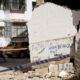 Crumbled concrete wall with rubble on a street, police tape across, and parked cars and a scooter in front of white apartment buildings.