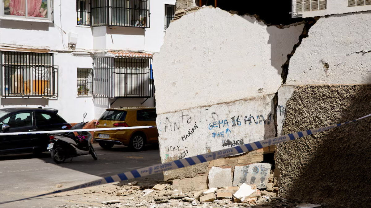 Crumbled concrete wall with rubble on a street, police tape across, and parked cars and a scooter in front of white apartment buildings.