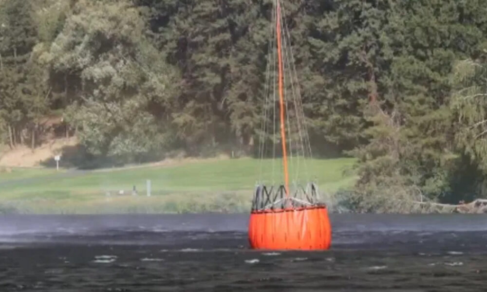 Large orange circular buoy floating on calm water with ropes extending upward, forested shore in background.